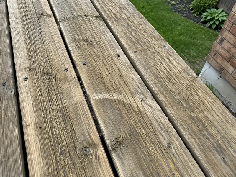 Close-up of grey pressure-treated deck boards with raised ridges along the grain, fuzzy lifted fibres and a pale arc across one board