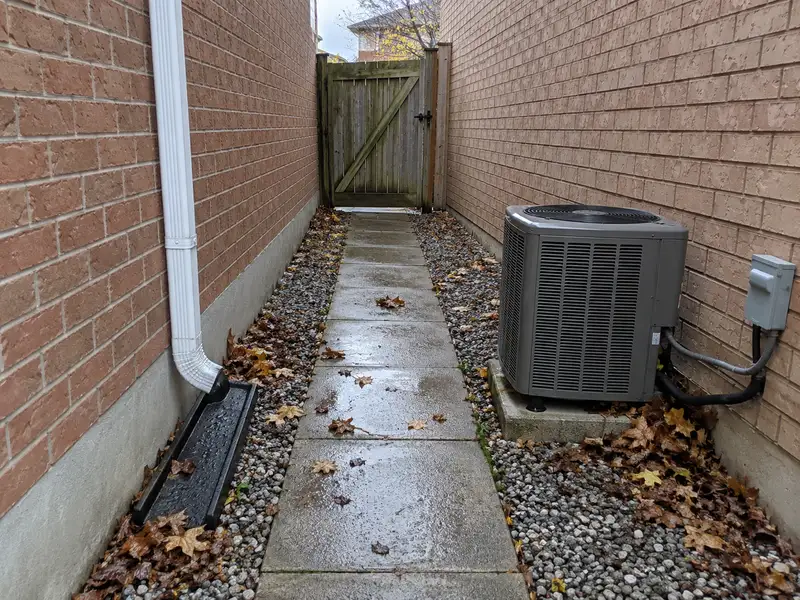 Narrow side yard between two brick houses, with a concrete slab path between gravel strips, an air conditioner unit, a downspout and a wooden gate at the far end
