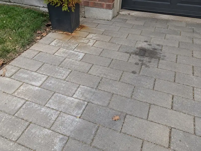 Grey concrete driveway pavers with orange rust marks beside a metal planter, a white powdery film on several pavers and a dark oil drip patch