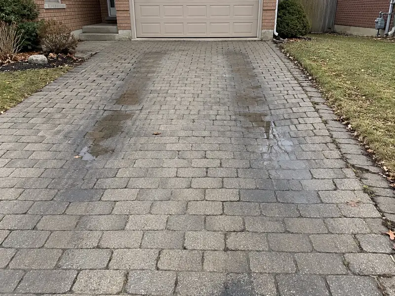 Older grey paver driveway with two dark, damp wheel tracks running toward the garage, rainwater pooled where the tracks have dipped, and weeds in the joints along the lawn edge