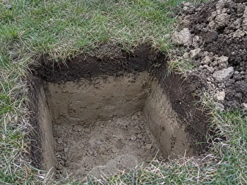 Square spade hole in a lawn showing a shallow band of dark topsoil over pale, dense clay subsoil